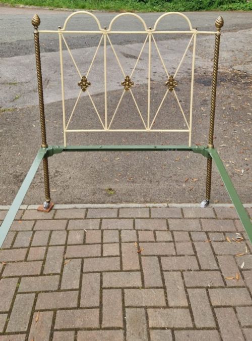 fig 1. A Victorian brass and iron single bedstead featuring twisted brass corner posts with urn finials, cream-painted arched metal framework with brass rosettes, and a green base.