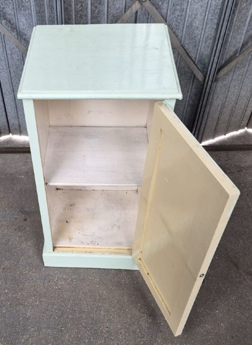 A narrow pale green painted Edwardian bedside cabinet featuring a single panelled door with a metal drop handle and a plinth base. View II