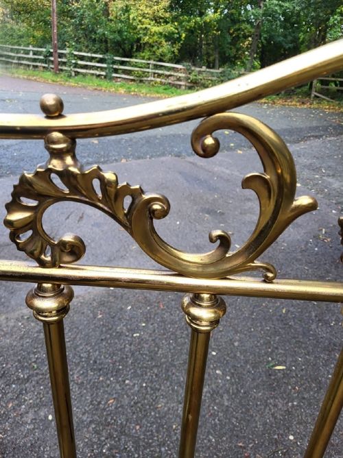 An antique Victorian brass king size bedstead featuring scrolled top rails, vertical spindles, floral cast cartouches, and metal castors. View II