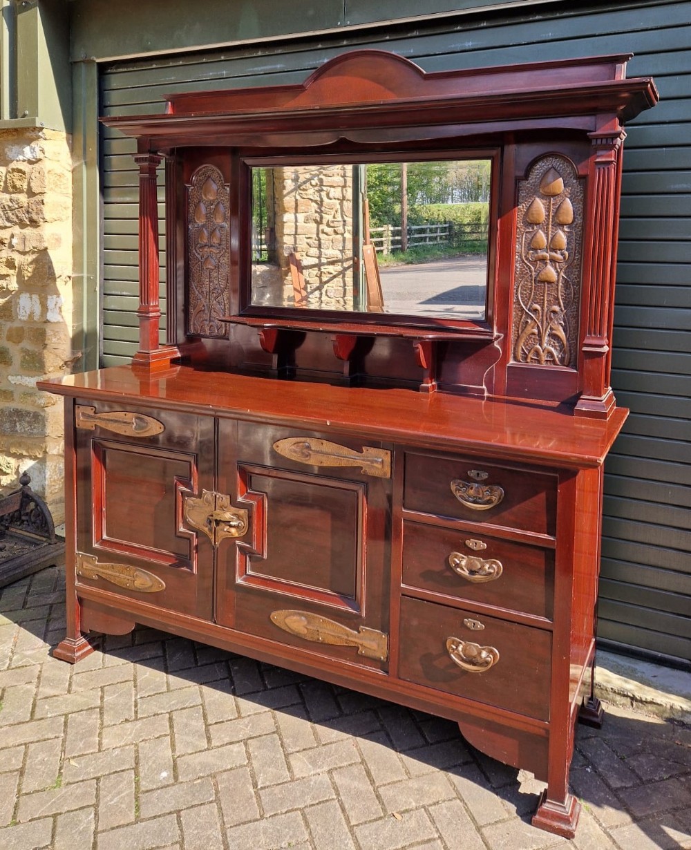 An Arts and Crafts mahogany sideboard featuring a mirrored upper structure, stylized repousse copper panels, and prominent brass strap hinges.
