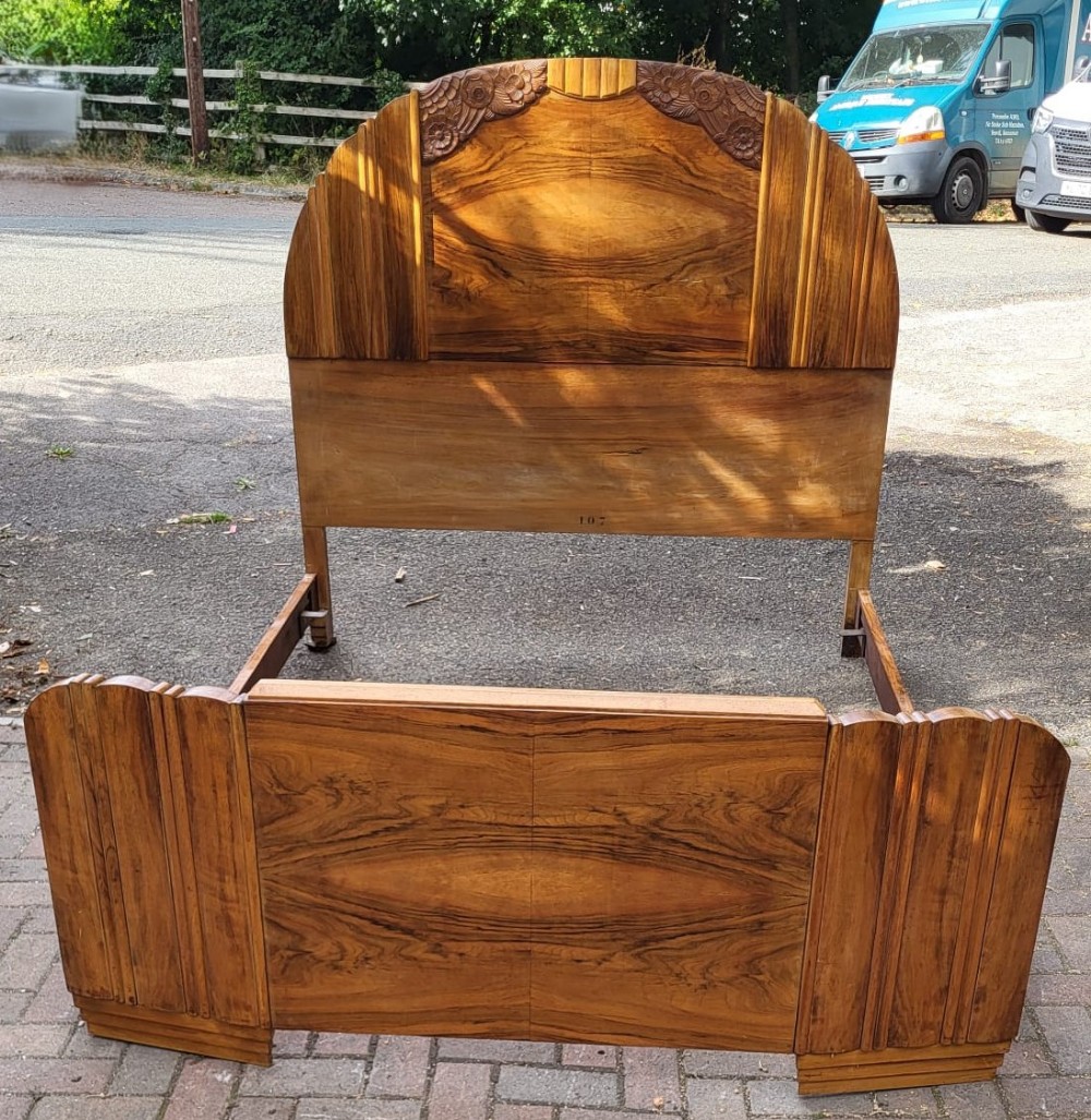 An Art Deco walnut bedstead featuring bookmatched timber veneers, a stepped arched headboard with carved floral details, and matching fluted side columns.