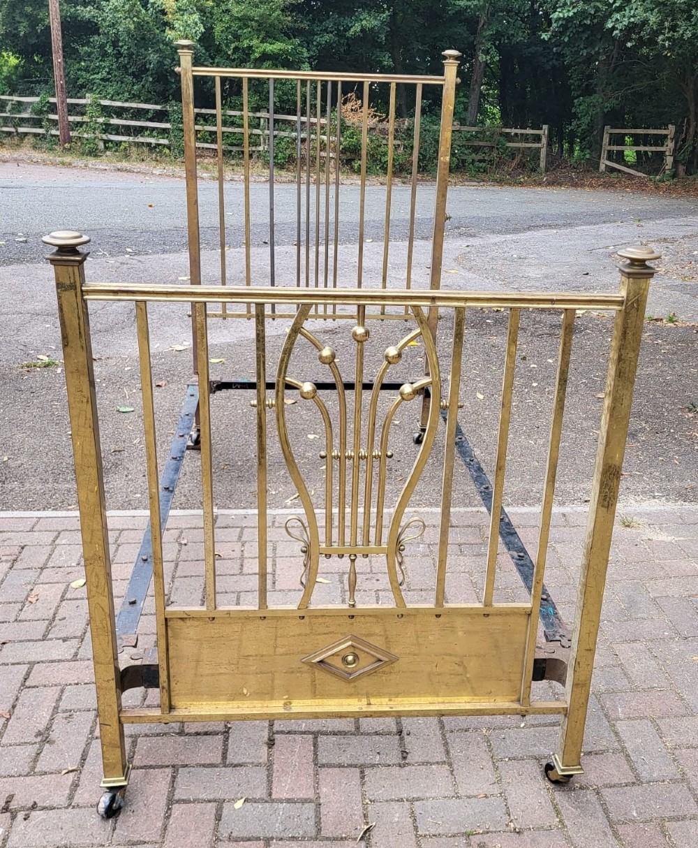 Late Victorian single brass bedstead featuring vertical spindle framework, a central stylised lyre motif on the footboard, square upright posts with finials, and castors.