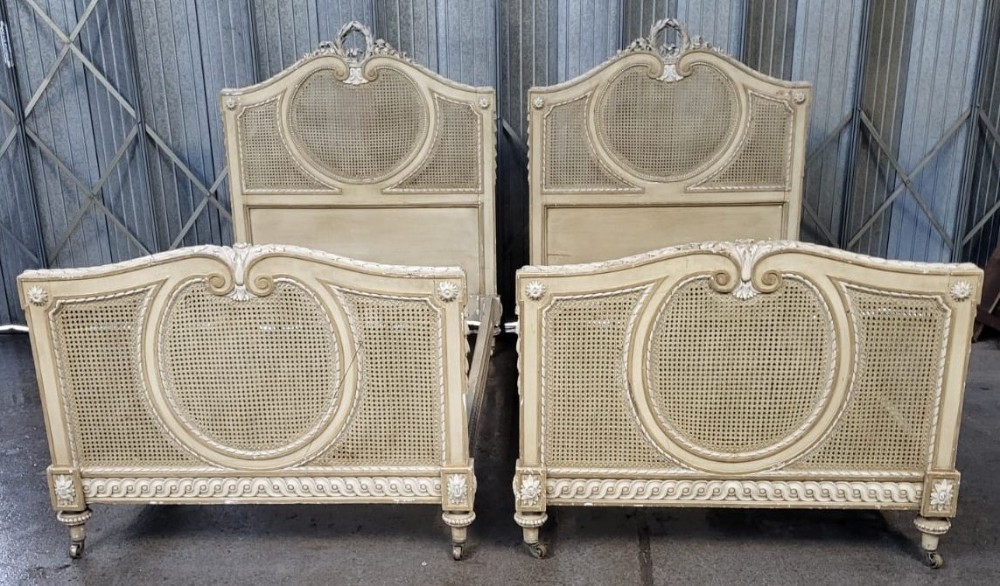 A pair of 19th-century French single bedsteads featuring cream painted frames with woven cane panels and ribbon cresting.