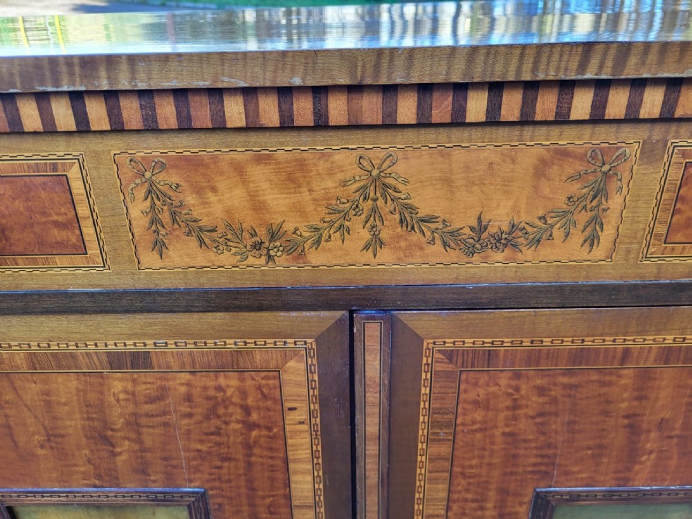 A golden satinwood side cabinet featuring painted figurative door panels, marquetry inlay swags, a top drawer with brass handles, and square tapered legs. View VII
