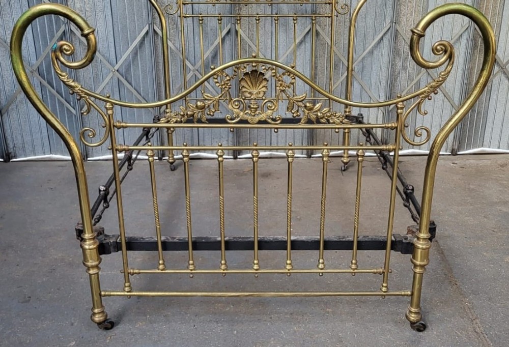 An ornate Victorian double brass bedstead featuring large outer scroll arms, a central shell casting, and vertical spindles on castors. View IV