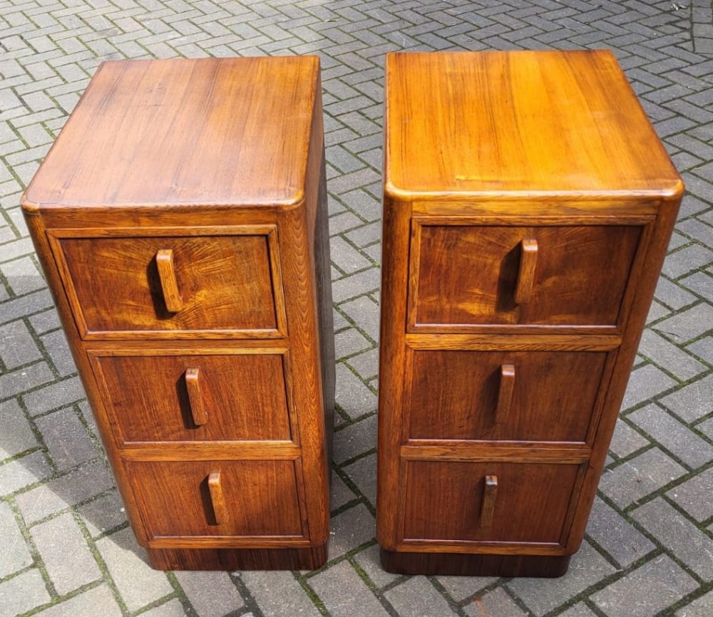 A matching pair of tall Art Deco oak bedside cabinets, each featuring three drawers with vertical wooden handles and curved upper corners.