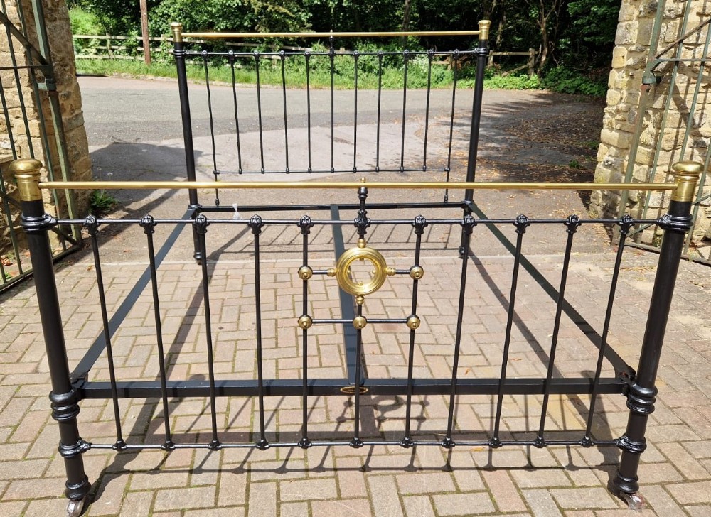 A Victorian super king size bedstead featuring a black painted cast iron frame with vertical spindles and polished brass top rails and central motif.