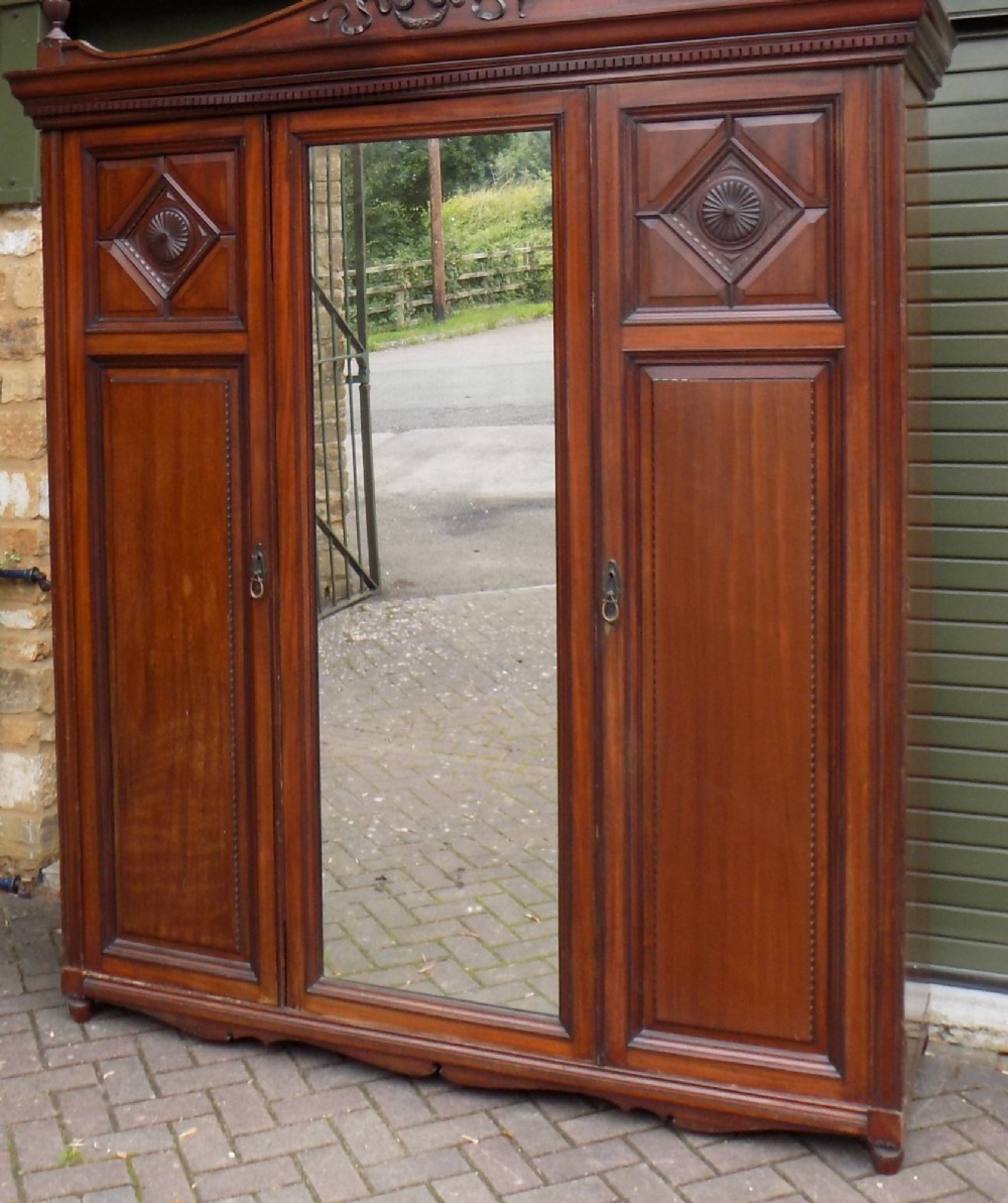 A late Victorian mahogany triple door wardrobe featuring a central full-length mirror, carved diamond relief panels, and a carved crest pediment.