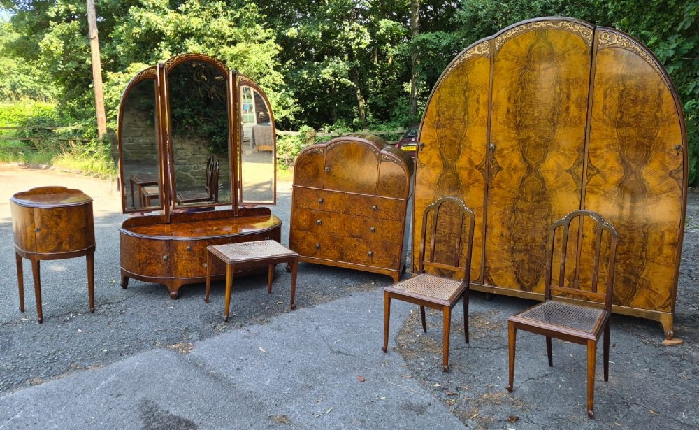 A seven-piece Art Deco bedroom suite crafted in burr walnut with arched tops, featuring a large wardrobe, triple-mirror dressing table, tallboy chest, bedside cupboard, stool, and two chairs.