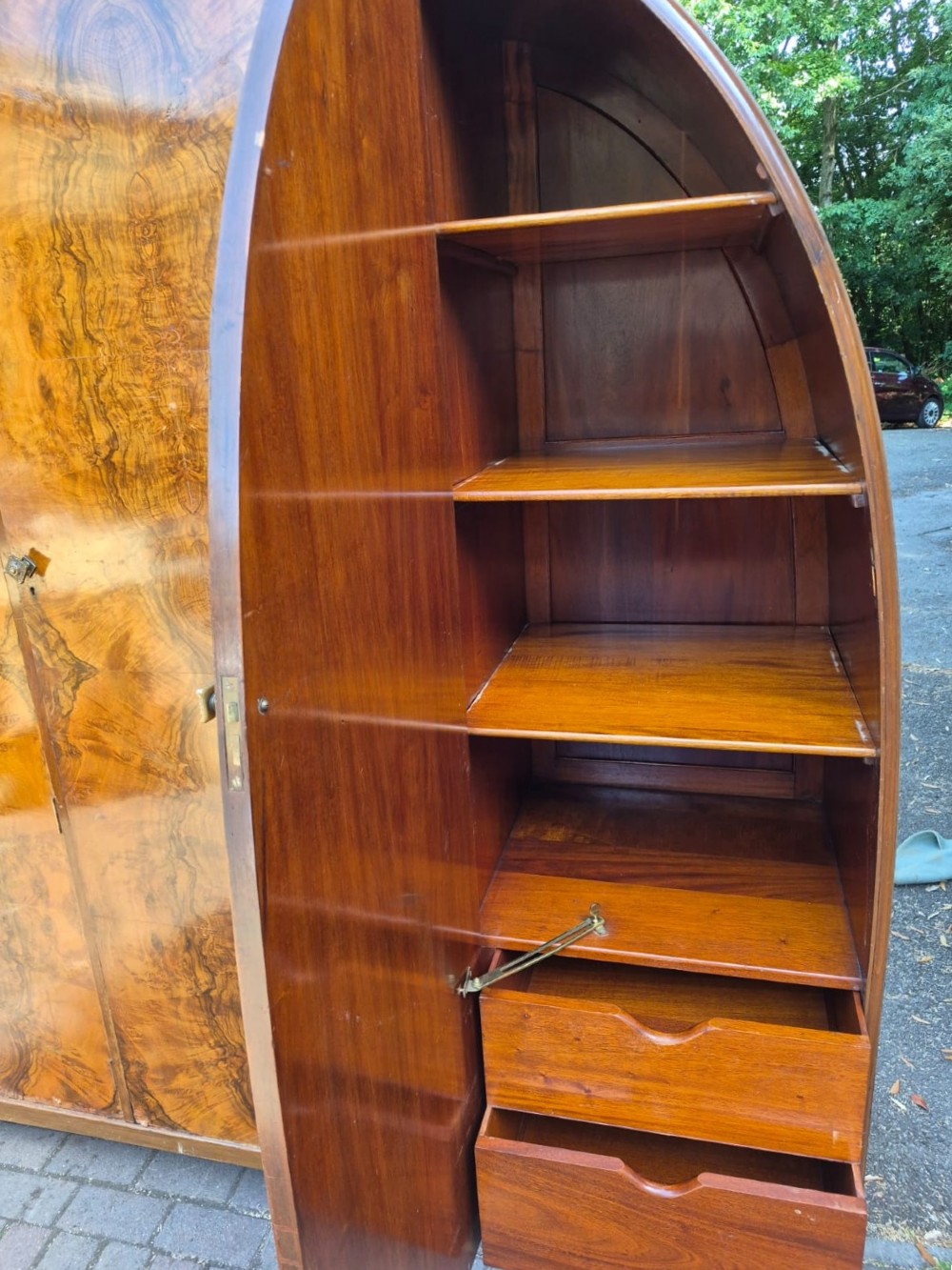 A seven-piece Art Deco bedroom suite crafted in burr walnut with arched tops, featuring a large wardrobe, triple-mirror dressing table, tallboy chest, bedside cupboard, stool, and two chairs. View III