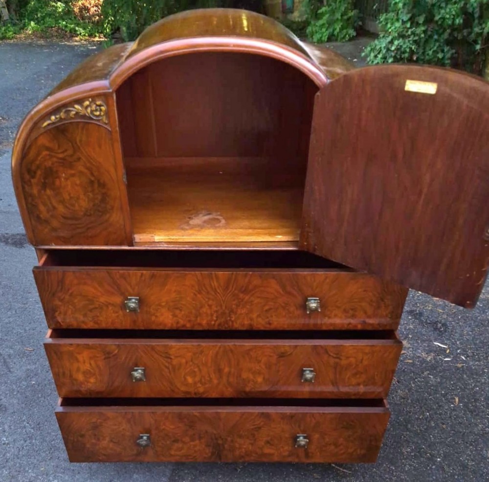 A seven-piece Art Deco bedroom suite crafted in burr walnut with arched tops, featuring a large wardrobe, triple-mirror dressing table, tallboy chest, bedside cupboard, stool, and two chairs. View V