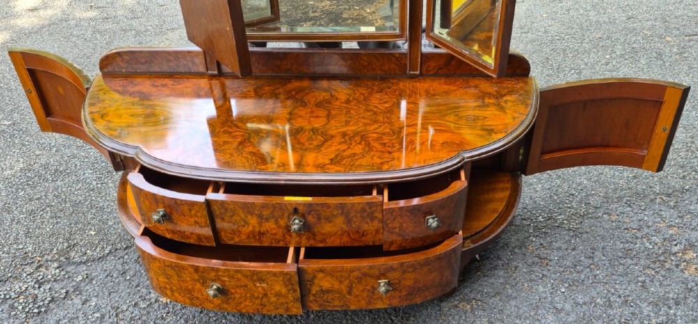 A seven-piece Art Deco bedroom suite crafted in burr walnut with arched tops, featuring a large wardrobe, triple-mirror dressing table, tallboy chest, bedside cupboard, stool, and two chairs. View X