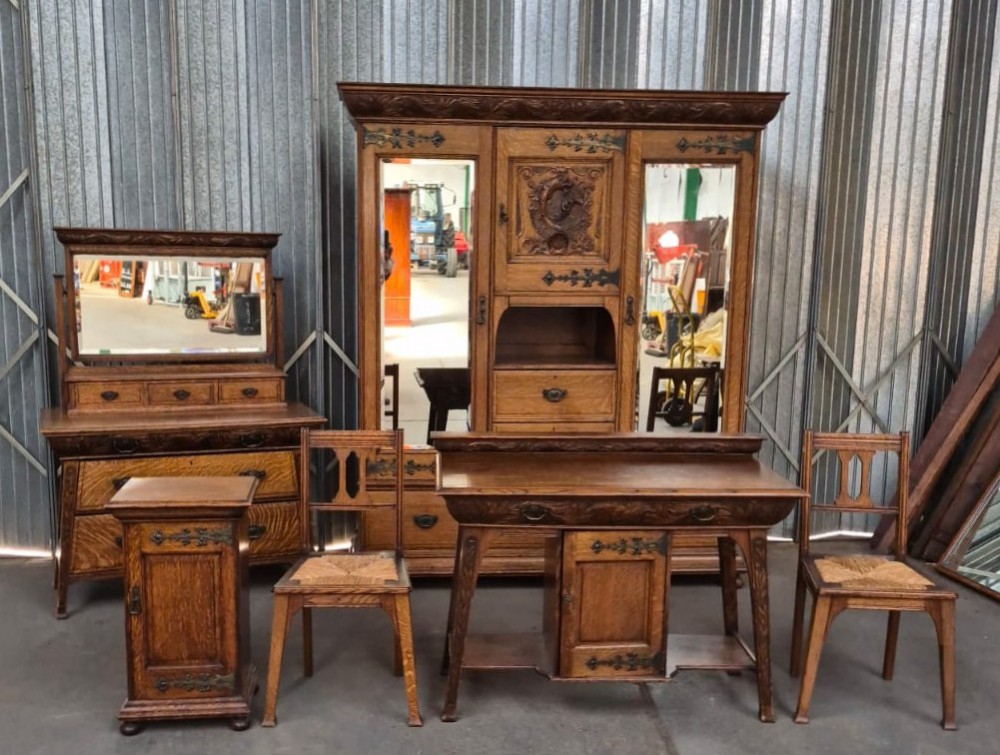 A six-piece golden oak Arts and Crafts bedroom suite including a central compactum wardrobe with mirrors, a dressing chest, a desk, a pot cupboard, and two rush-seat chairs.