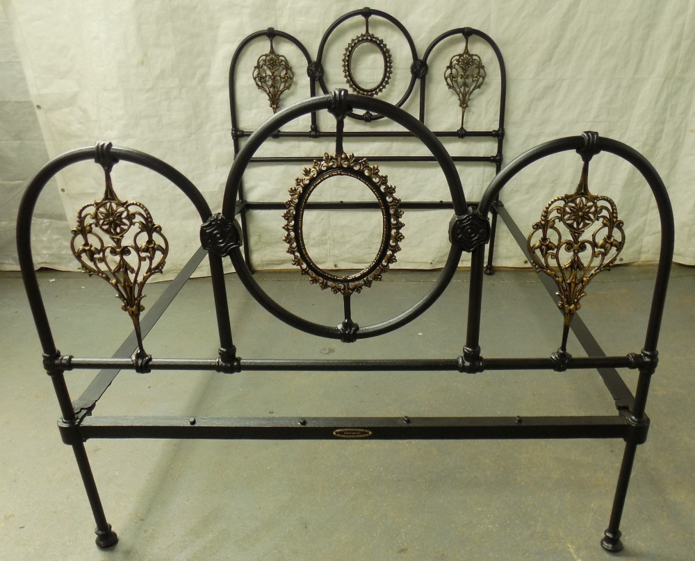 A Victorian double iron bedstead featuring a black tubular frame with arched top rails and central circular medallions with gold filigree detail. View III