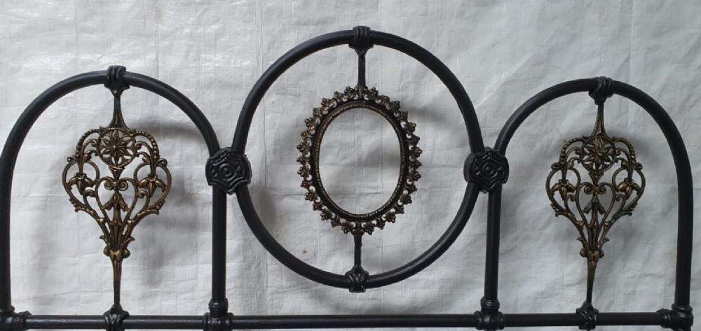 A Victorian double iron bedstead featuring a black tubular frame with arched top rails and central circular medallions with gold filigree detail. View IV