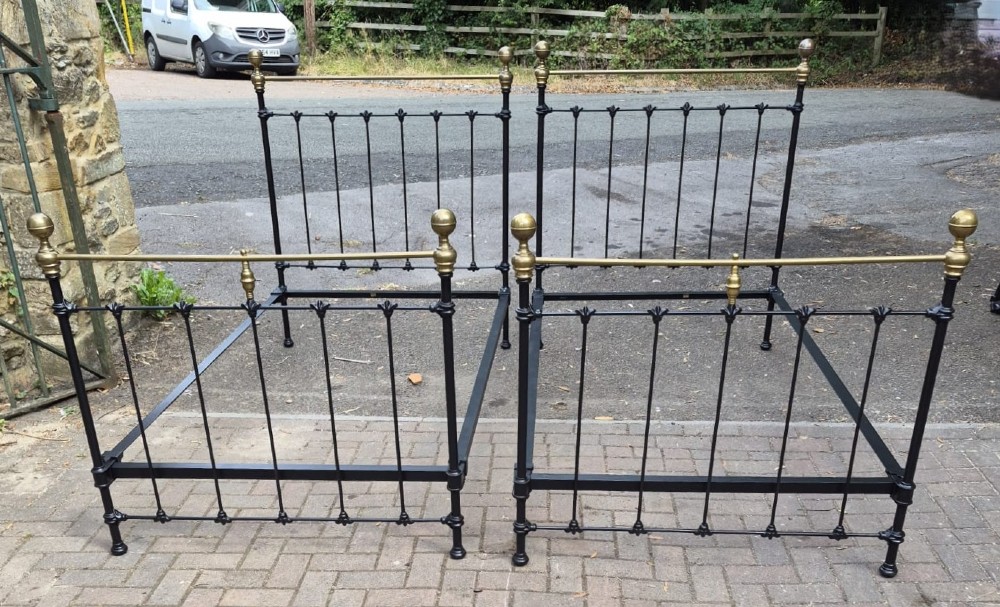 A matching pair of Victorian black iron single bedsteads featuring vertical spindles, cast floral embellishments, and polished brass top rails with spherical finials.