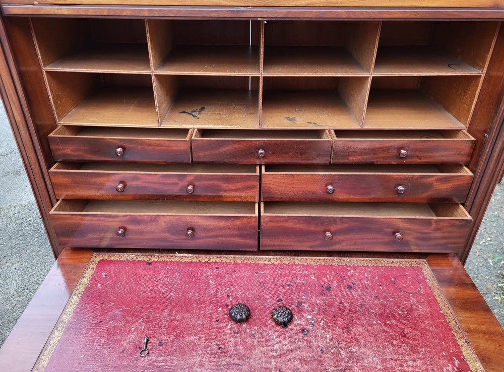 A 19th-century flame mahogany secretaire a abattant featuring carved scrollwork, diamond veneer patterns, lower cupboards with rosette medallions, and a brass gallery top. View IV