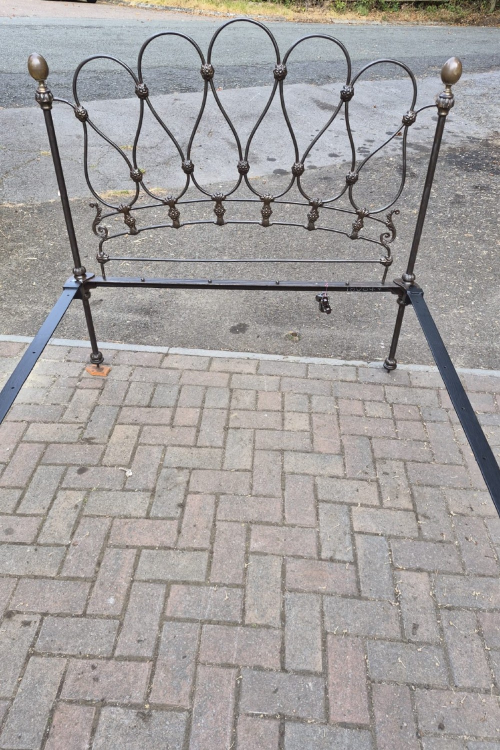 A Victorian single bedstead crafted from black iron with intricate curved scrollwork loops and brass urn finials on the corner posts. View II