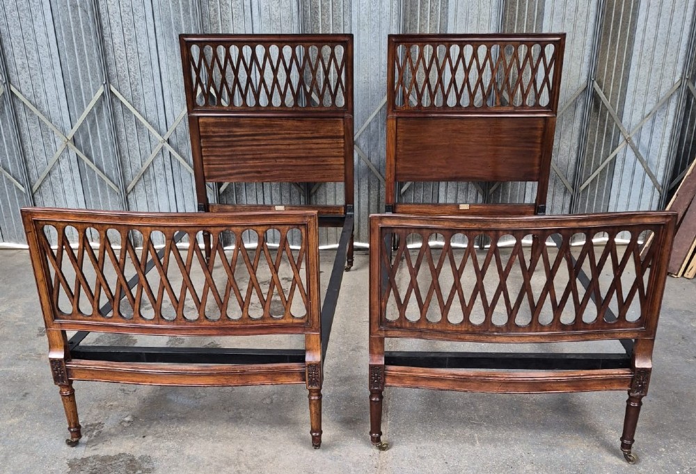 A pair of Edwardian mahogany single bed frames featuring pierced lattice upper panels, flame mahogany lower panels, turned legs, and brass castors.