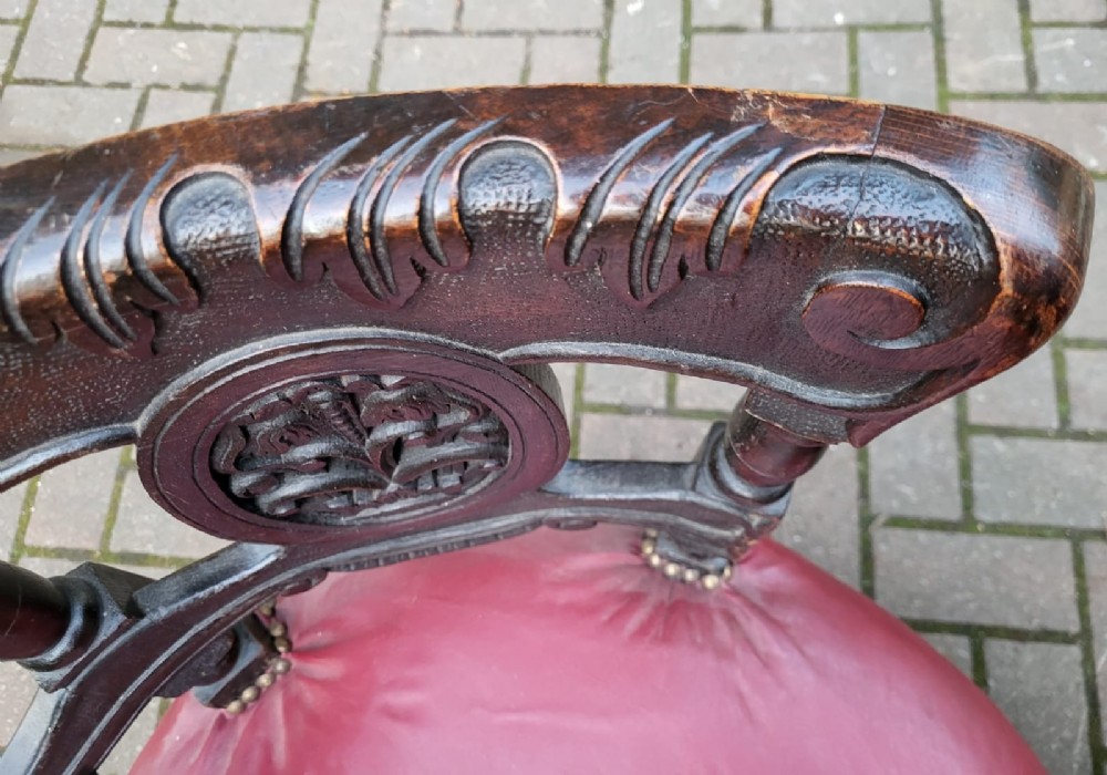 A Victorian carved dark oak Burgomaster chair with a curved back rail, carved oval panels, and a red leather circular seat with brass stud details. View III
