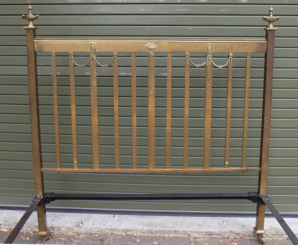 An antique Victorian brass bedstead with decorative urn finials, vertical spindles, ribbon swags, and castors. View II