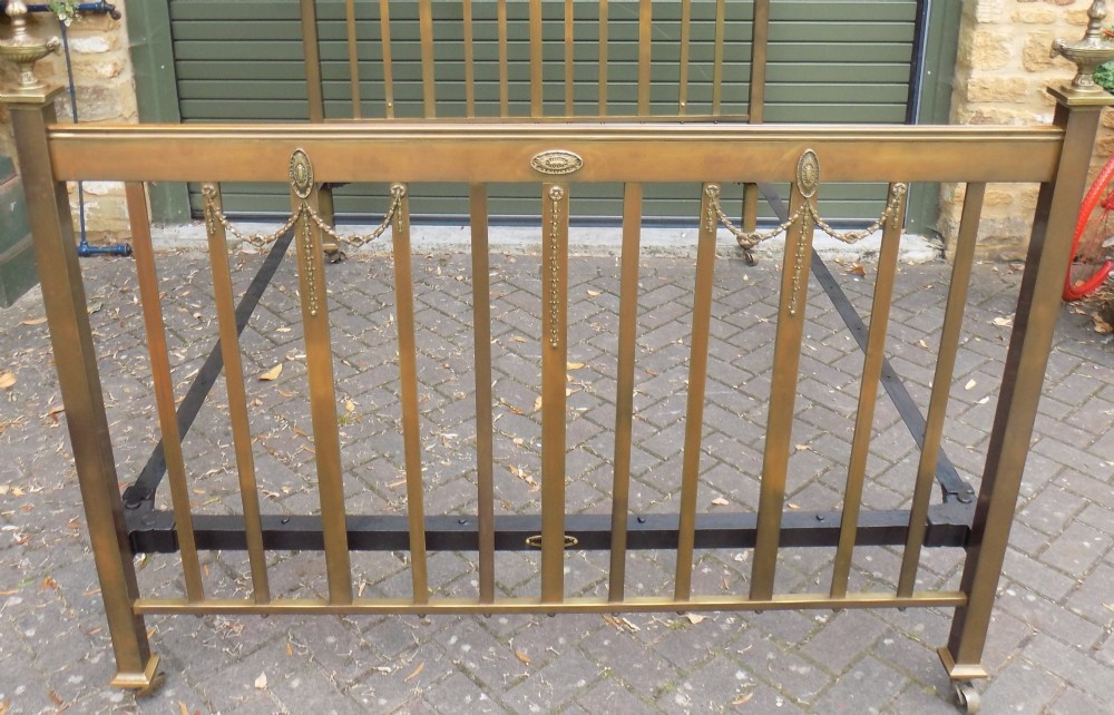 An antique Victorian brass bedstead with decorative urn finials, vertical spindles, ribbon swags, and castors. View III