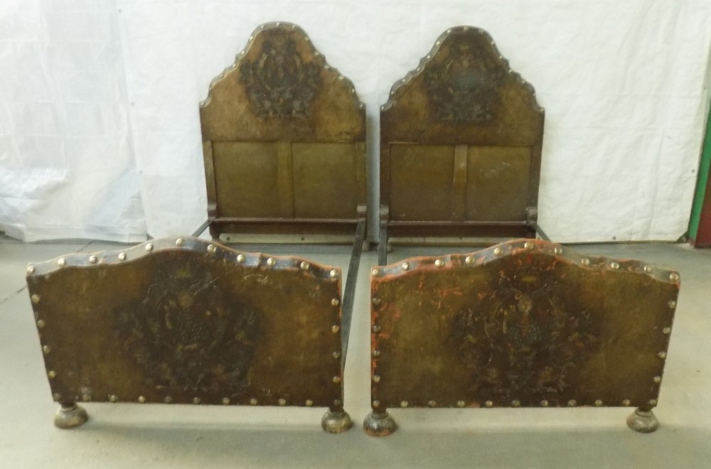 A pair of matching single bedsteads upholstered in aged brown hand-painted leather with brass stud trimming and arched tops.