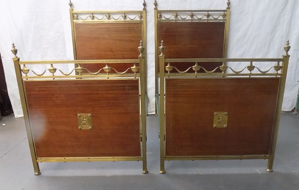 A pair of late Victorian brass-framed twin bedsteads featuring polished brass rails, top drapery swags, urn finials, and central mahogany timber panels with decorative brass plaques. View III