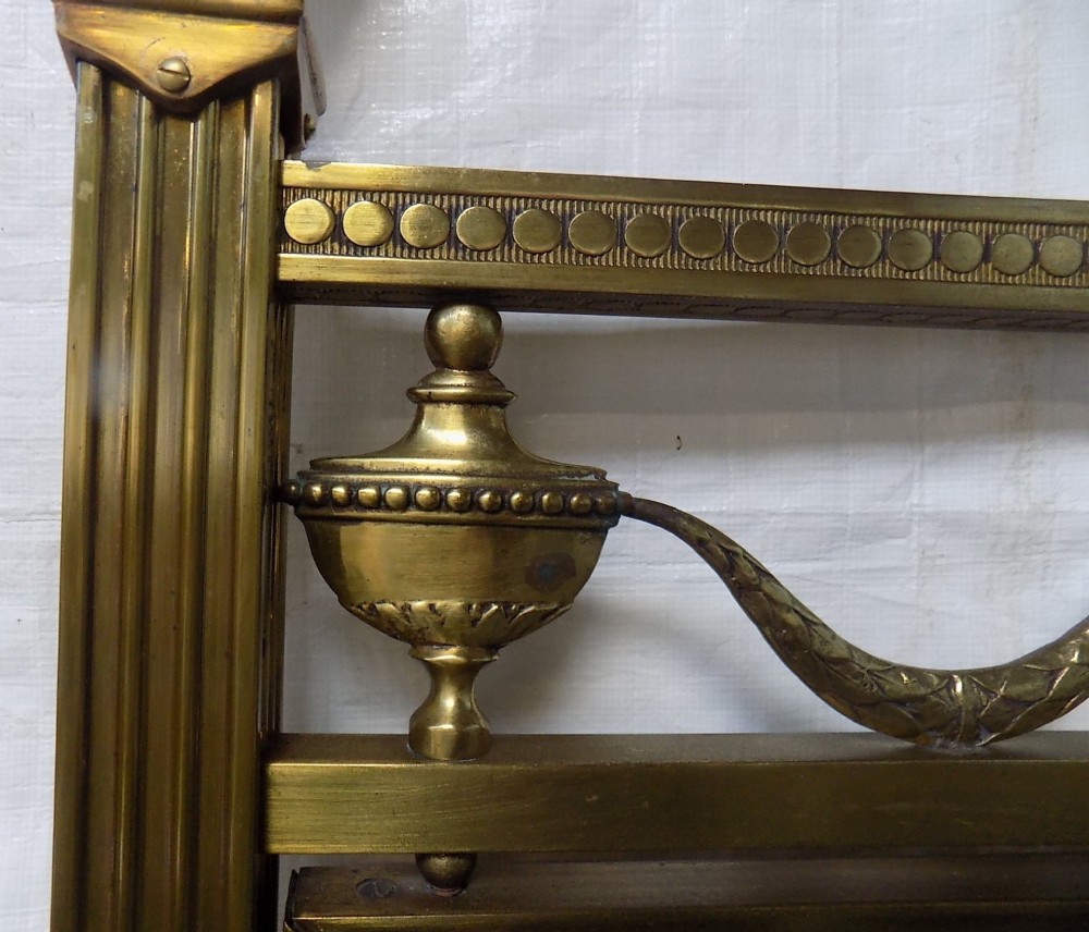 A pair of late Victorian brass-framed twin bedsteads featuring polished brass rails, top drapery swags, urn finials, and central mahogany timber panels with decorative brass plaques. View V