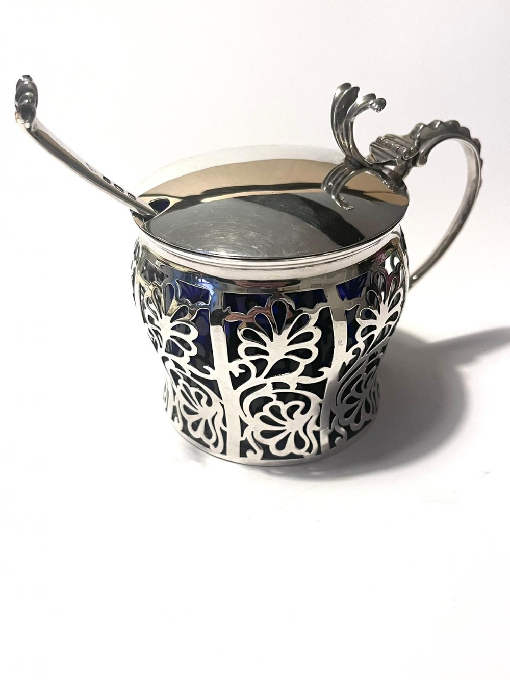 An Art Deco sterling silver jam pot featuring pierced floral scrollwork, a hinged lid, a matching spoon, and a dark cobalt blue glass liner.