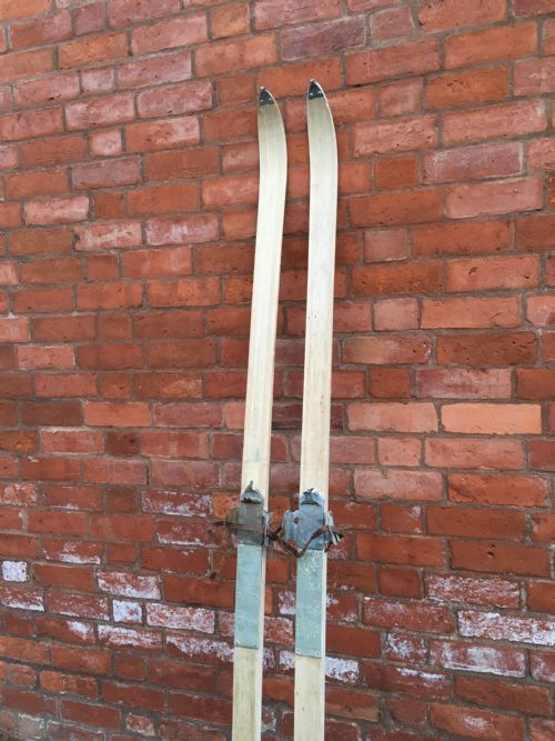 fig 2. A pair of tall light-coloured wooden skis featuring metal bindings and leather straps positioned vertically.