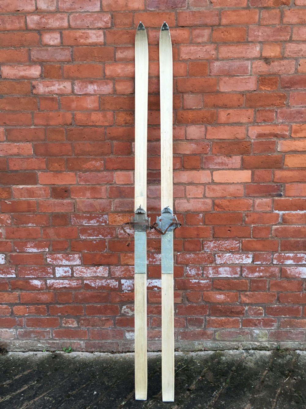 A pair of tall light-coloured wooden skis featuring metal bindings and leather straps positioned vertically.