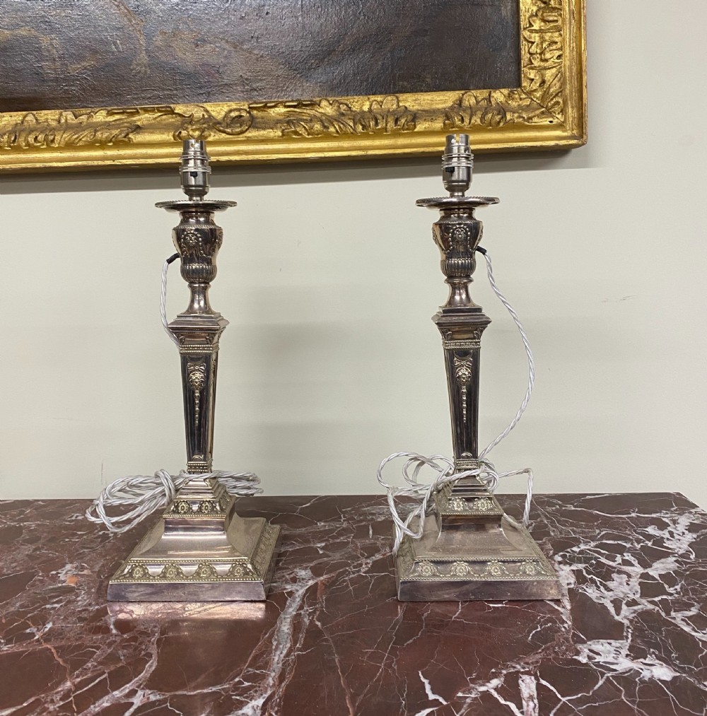 A pair of 19th-century Neoclassical silver-plated table lamps featuring tapered square column shafts and classical urn-shaped sockets on decorated square bases.