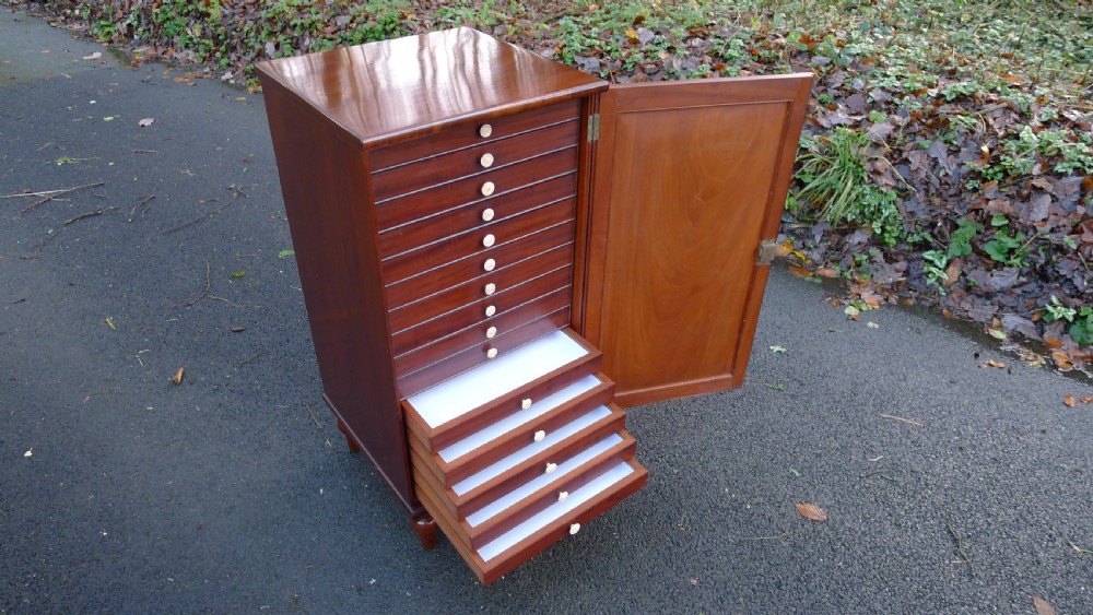 A nineteenth-century mahogany collectors cabinet featuring an open panelled door revealing multiple shallow drawers with light-coloured knob handles.