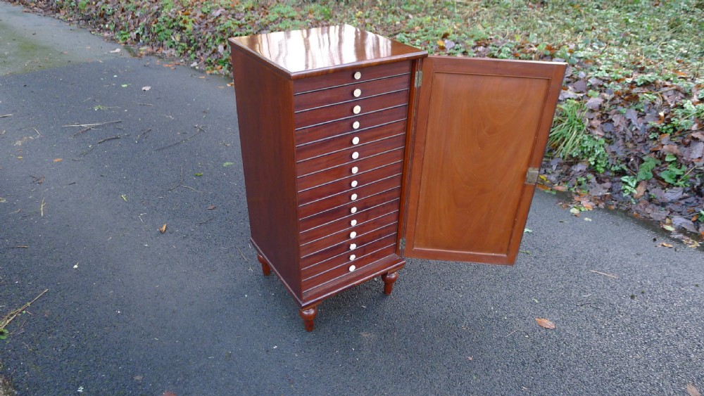 A nineteenth-century mahogany collectors cabinet featuring an open panelled door revealing multiple shallow drawers with light-coloured knob handles. View IV