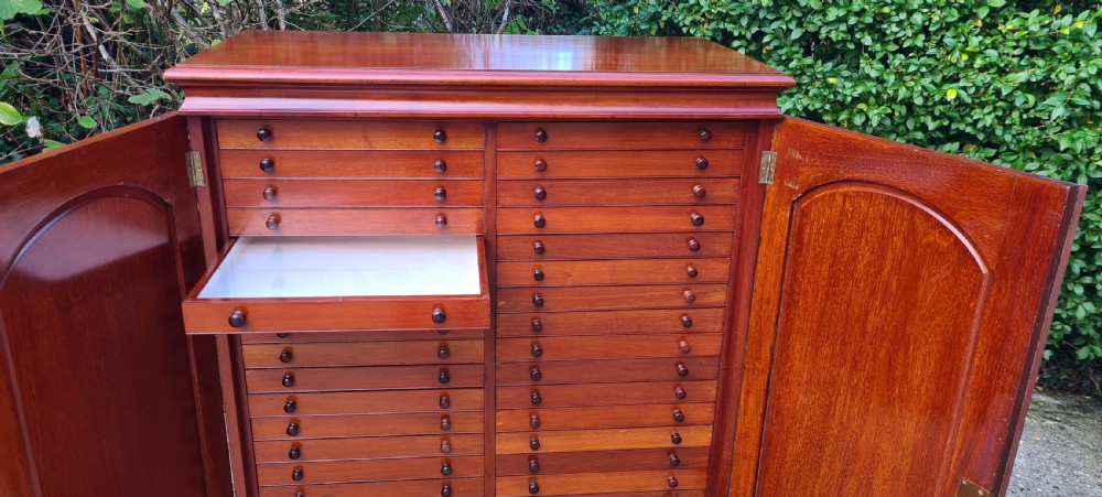 An Edwardian mahogany collectors cabinet with double doors open to reveal forty shallow drawers with small turned wooden knobs, featuring one drawer pulled forward displaying a glass cover. View XV