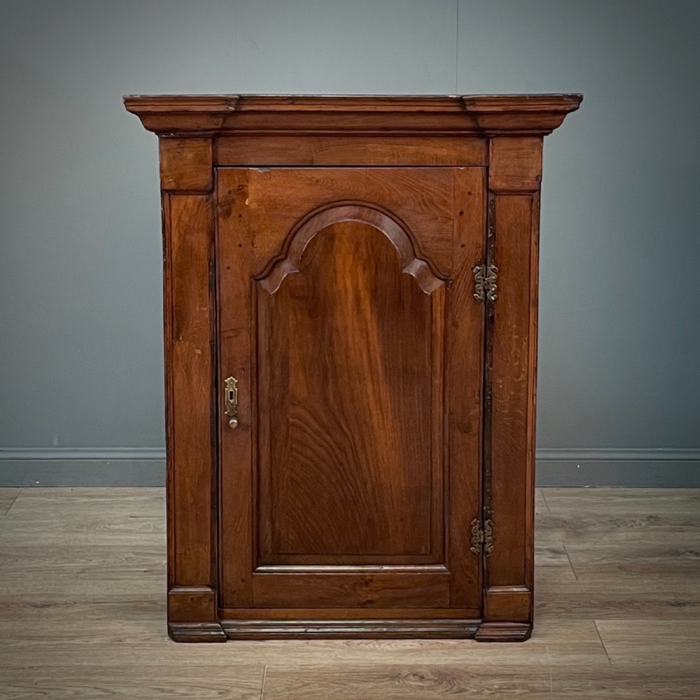 An antique Georgian oak hanging corner cabinet featuring an arched panel door, moulded top cornice, and brass fittings. View II