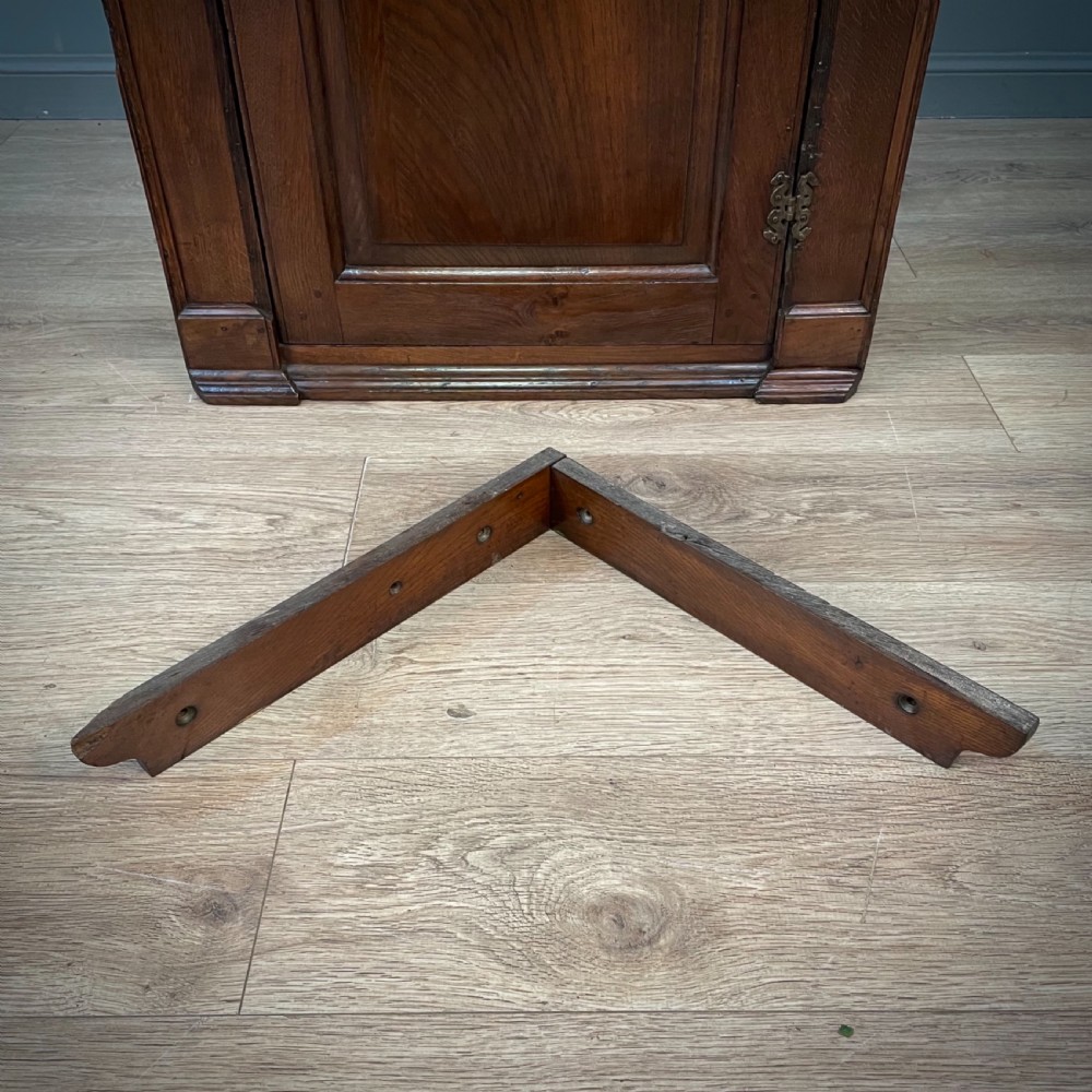 An antique Georgian oak hanging corner cabinet featuring an arched panel door, moulded top cornice, and brass fittings. View X