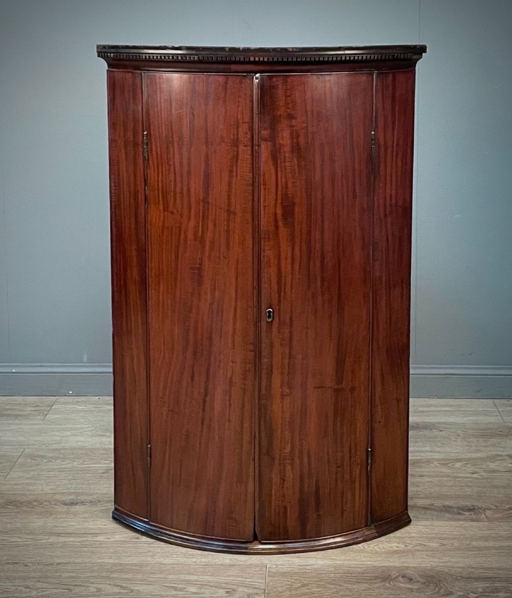 A Georgian mahogany bow-front hanging corner cabinet featuring double doors with flame timber grain, brass escutcheon, and a dentil-moulded top cornice.