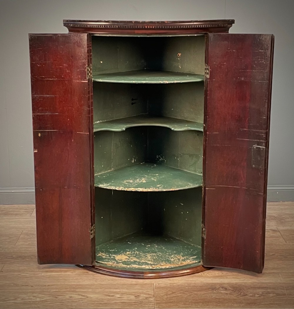 A Georgian mahogany bow-front hanging corner cabinet featuring double doors with flame timber grain, brass escutcheon, and a dentil-moulded top cornice. View II