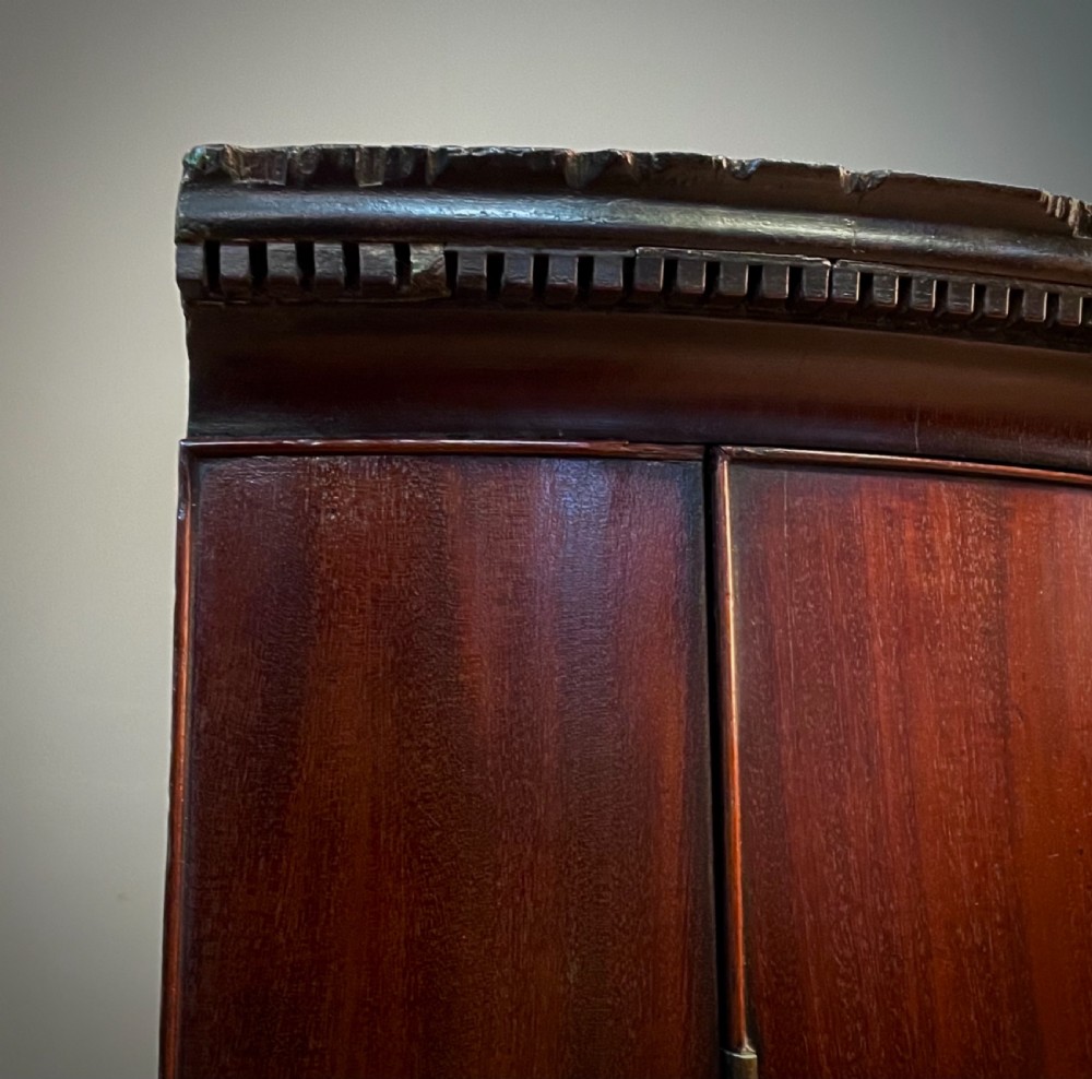 A Georgian mahogany bow-front hanging corner cabinet featuring double doors with flame timber grain, brass escutcheon, and a dentil-moulded top cornice. View IV