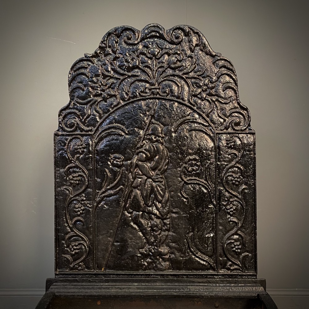 A black cast and wrought iron fire basket featuring a relief-decorated fireback, slatted log holder, scrolled iron side supports, and polished brass ball finials. View II