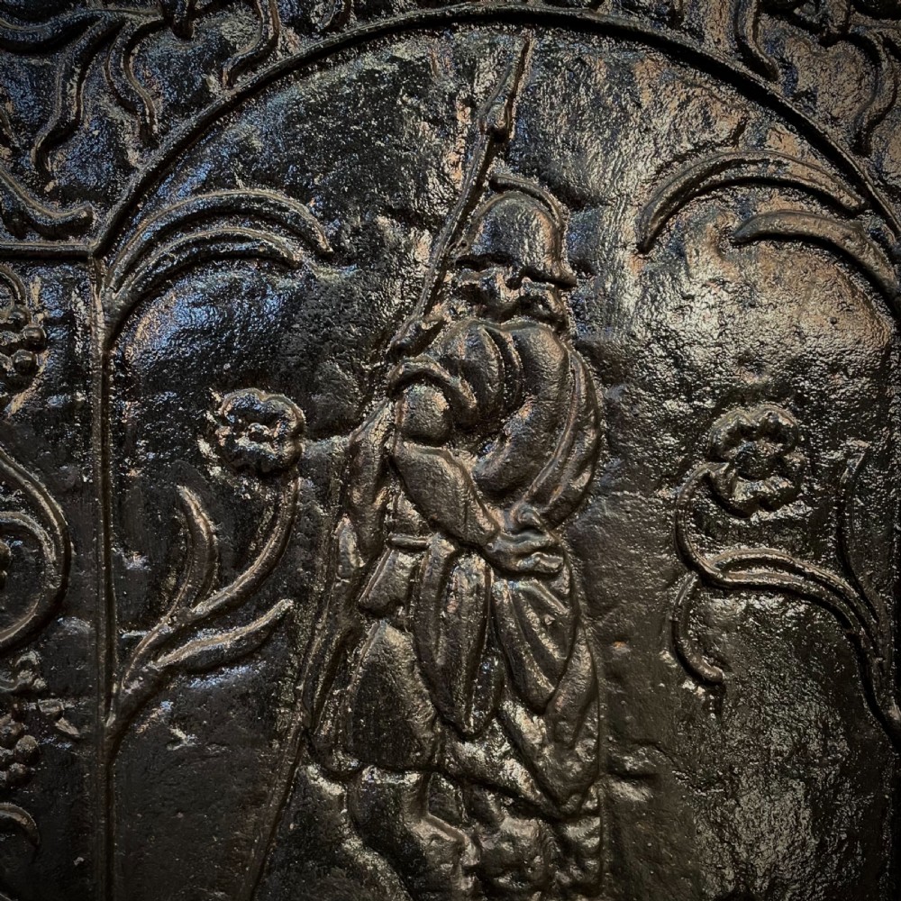 A black cast and wrought iron fire basket featuring a relief-decorated fireback, slatted log holder, scrolled iron side supports, and polished brass ball finials. View VII