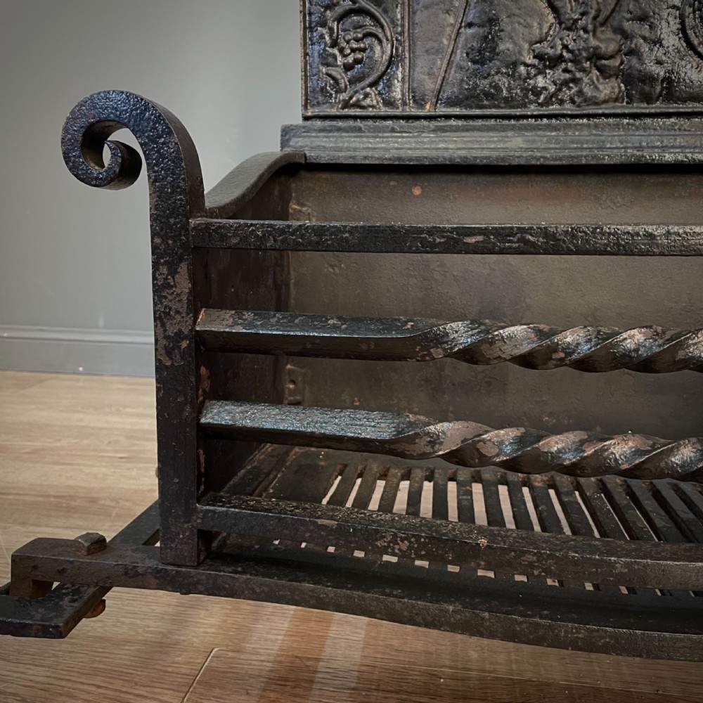 A black cast and wrought iron fire basket featuring a relief-decorated fireback, slatted log holder, scrolled iron side supports, and polished brass ball finials. View VIII