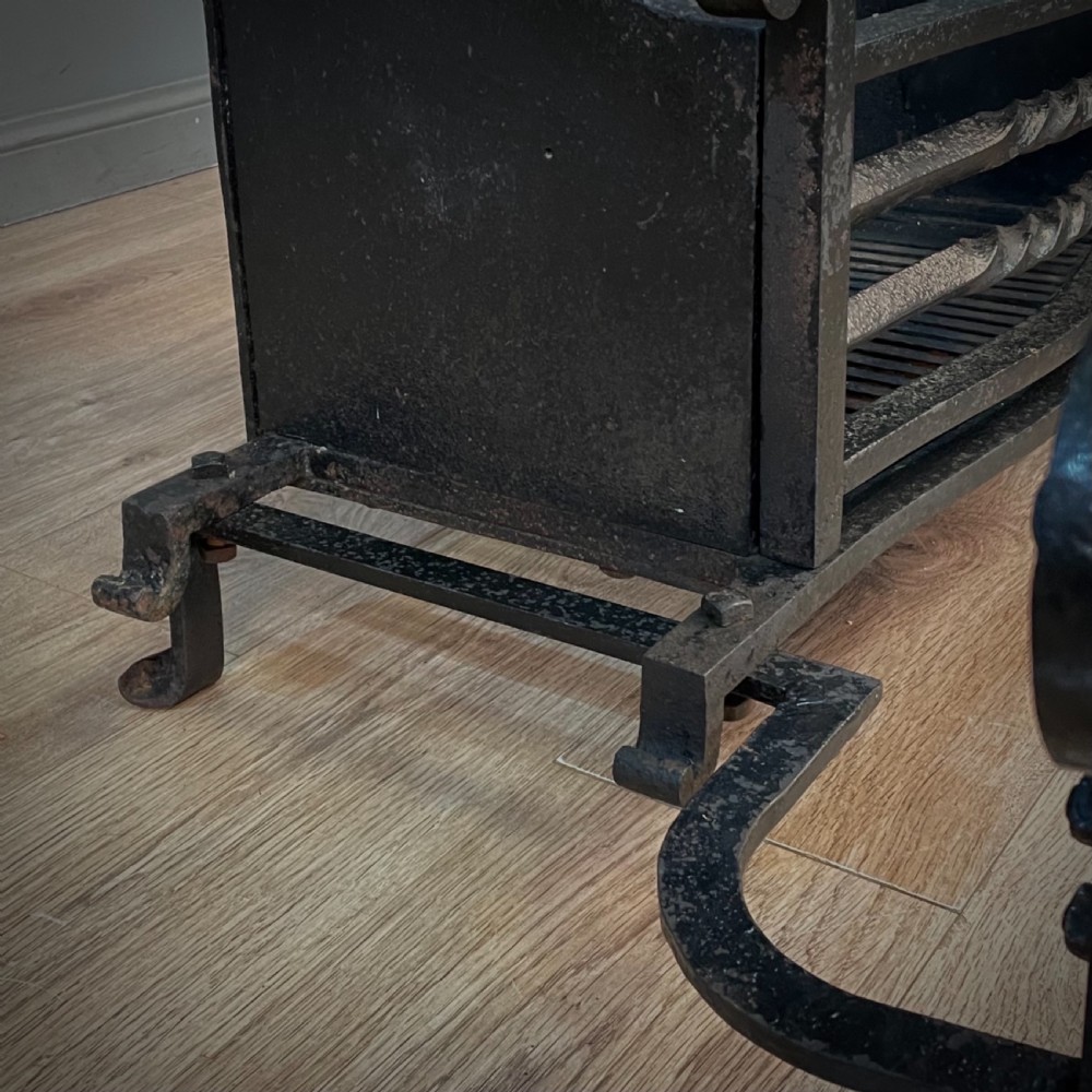 A black cast and wrought iron fire basket featuring a relief-decorated fireback, slatted log holder, scrolled iron side supports, and polished brass ball finials. View XII