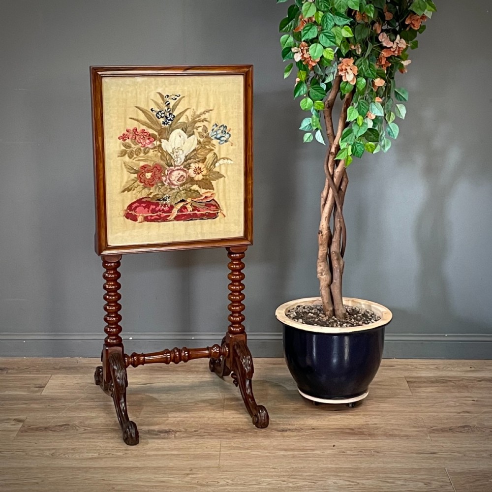 A Victorian mahogany fire screen featuring bobbin-turned uprights, carved feet, and a framed needlework panel displaying a floral bouquet.
