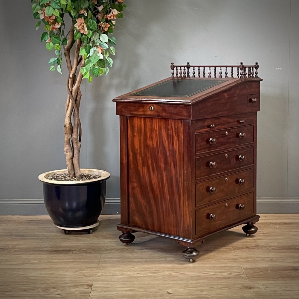A William IV mahogany Davenport desk featuring a sloped leather writing slope, turned spindle gallery top, side drawers, and brass castors.
