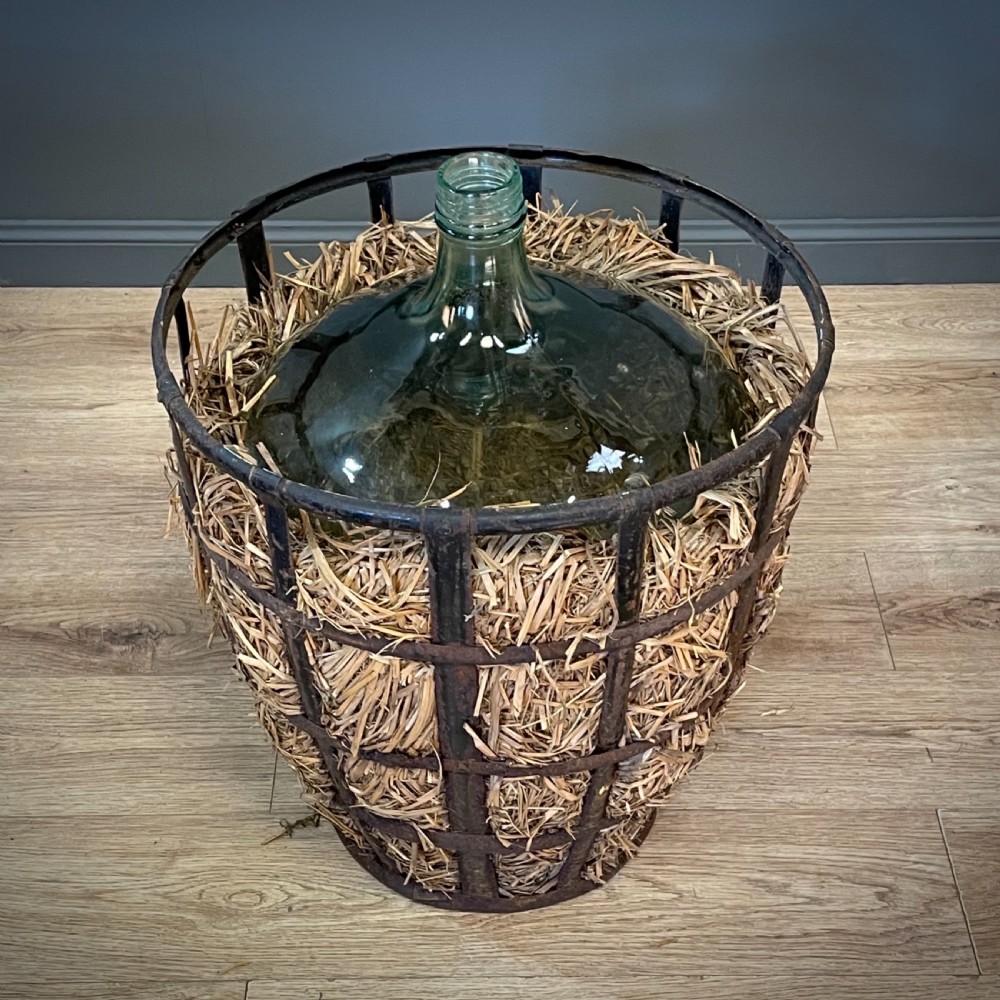 A large green glass demijohn bottle resting inside a natural straw-lined iron basket frame. View II