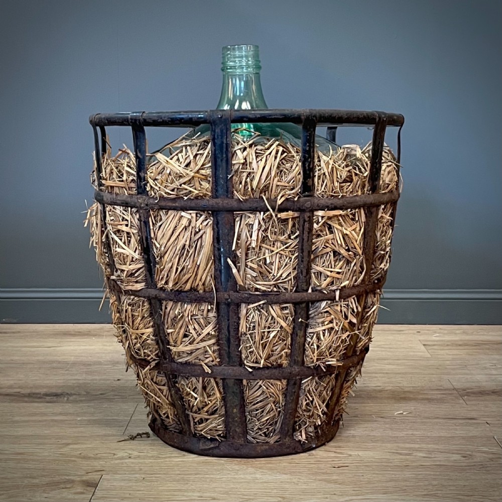 A large green glass demijohn bottle resting inside a natural straw-lined iron basket frame. View III