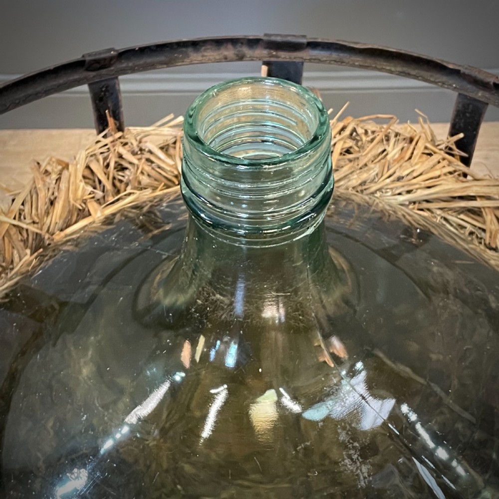 A large green glass demijohn bottle resting inside a natural straw-lined iron basket frame. View IV